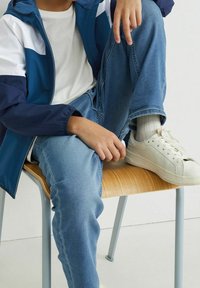 Námořnická a bílá dvoubarevná bunda, bílý tričko, modré džíny a bílé tenisky. Dřevěná židle s modrými kovovými nohami.