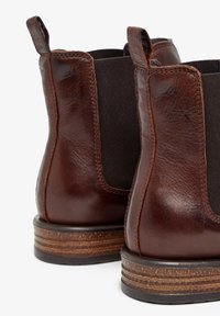 Botas de cuero marrón hasta el tobillo con paneles laterales elásticos, punta redondeada y tacón de madera apilada. Detalle de costura notable a lo largo de los bordes.