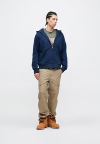 Tamsiai mėlynas su užtrauktuku džemperis užsegamas virš alyvuogių spalvos marškinėlių, derinamas su smėlio spalvos krovinių kelnėmis ir rudos darbo batais. Ypatybės: kišenė ant pilvo ir reguliuojama gaubtas.