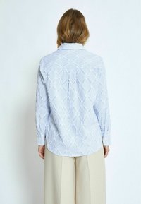 Camicia button-up azzurro chiaro con righe verticali e un motivo geometrico. Presenta maniche lunghe e un orlo curvo. Tessuto texturizzato.