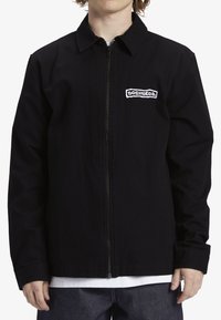 Chaqueta negra con cremallera fabricada en un material duradero, presenta un cuello tipo solapa y un logo bordado en el lado izquierdo del pecho. Mangas largas, diseño sencillo.