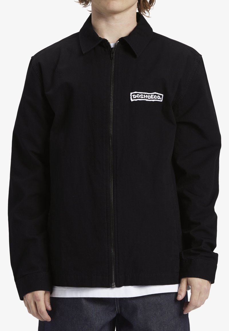 Chaqueta negra con cremallera fabricada en un material duradero, presenta un cuello tipo solapa y un logo bordado en el lado izquierdo del pecho. Mangas largas, diseño sencillo.