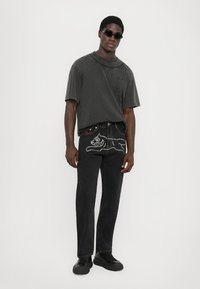 Mørkegrå oversized t-skjorte med strukturert rundhals, sammen med svarte jeans med sølvnitter formet som en katt og rød knappeakcent.