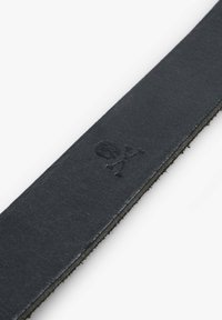 Correa de cuero negro con una textura suave, que presenta un detalle en relieve de calavera y tibias cruzadas. Forma rectangular, diseño estructurado y minimalista.