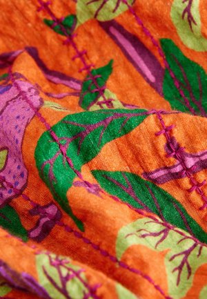 Leuchtend farbiger Stoff mit orangefarbenem Hintergrund, grünen Blättern und lila Akzenten, sichtbaren Nähten in pinkfarbenem Faden und weicher Textur.