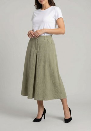 Femme portant une chemise blanche à manches courtes, un pantalon midi ample taille haute couleur vert olive et des talons hauts noirs, debout sur un sol gris.