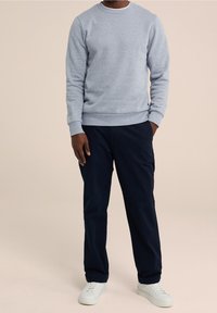 Man i ljusgrå långärmad sweatshirt, mörkblå byxor och vita sneakers, står med en hand i fickan mot en beige bakgrund.