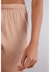 Satin loungewear broek in een lichtbeige kleur. Voorzien van een elastische tailleband en een gladde textuur. Huid is gedeeltelijk zichtbaar aan de rand.