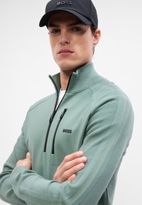 Hellgrüner Reißverschluss-Sweatshirt mit strukturiertem Stoff, schwarzem Reißverschluss und Logo. Das Model trägt eine schwarze Mütze; der Hintergrund ist schlicht und untexturiert.