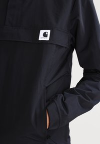 Sorte letvægtsjakke med en frontlomme og Carhartt-logo på en hvid label. Har høj krave og manchetter.