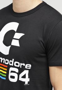 Camiseta de algodón negra con un gran logotipo blanco de "C" y el texto "Commodore 64" en blanco y acentos de colores. Mangas cortas, cuello redondo.