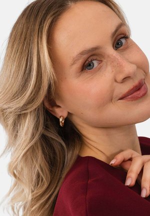 Frau mit blondem Haar und blauen Augen, die goldene Creolen und ein bordeauxrotes Oberteil trägt, lächelt sanft mit der Hand in der Nähe ihres Halses.