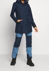 Parka imperméable bleu marine avec capuche, deux poches avant, associée à un pantalon bicolore bleu et marine et des bottes noires. Texture lisse.