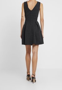 Svart ärmlös klänning med V-formad rygg, plisserad kjol och strukturerad tyg. Buren med svarta högklackade skor, som visar upp släta ben.