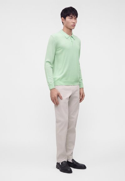 Homme debout portant un polo vert clair à manches longues, un pantalon beige et des mocassins noirs sur un fond blanc uni.