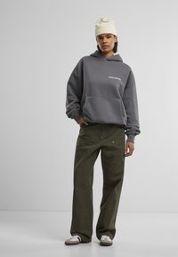 Grijze hooded sweatshirt met een voorkruiszak, gecombineerd met olijfgroene wijde broek. De outfit wordt aangevuld met een crèmekleurige gebreide beanie op het hoofd.