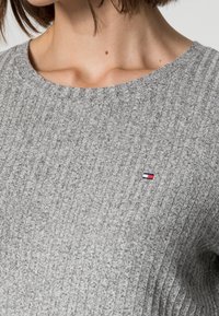 Grå ribbad tröja med rund halsringning, med en liten broderad logotyp i rött, vitt och blått på vänster bröst. Strukturerad tyg.