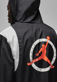 Giacca bianca e nera con cappuccio, caratterizzata da un grande logo arancione di Air Jordan sul retro. Tessuto liscio e lucido con un blocco di colori ben definito.