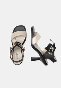 Black and beige heeled sandals featuring a wide strap, skinny heel, and decorative elastic with a button accent. Smooth leather texture.