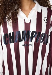 Close-up van een kastanjebruin en wit gestreept shirt met zwarte "CHAMPION" tekst, kleine "GAME ON" tekst en een voetbalgrafiek op de borst.