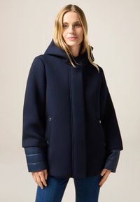 Cappotto con cappuccio blu navy in materiale morbido, dotato di bottoni a pressione, tasche laterali e polsini blu trapuntati. Design semplice e strutturato.