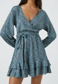 Woman wearing a blue floral wrap dress with long puffed sleeves, a tied waist belt, and a tiered ruffled hem.