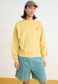 Sweatshirt jaune à col rond avec des poignets côtelés, doté d'une petite étiquette de logo. Associé à un short en velours côtelé bleu clair, coupe ample, longueur au-dessus du genou.