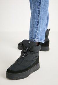 Botas de estilo puffers en color negro con una superficie texturizada, que cuentan con un upper acolchado, cierre con cordones y una suela gruesa y resistente.