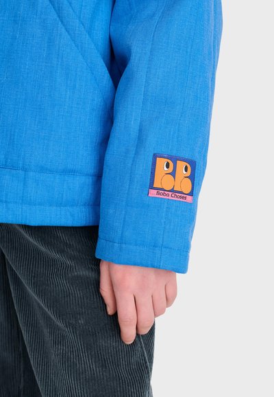 Main d'enfant près de la manche d'une veste bleue avec un petit patch montrant deux visages oranges et le texte "Bobo Choses" sur un pantalon en velours côtelé gris.