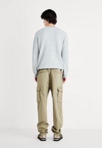 Lyseblå ribbestrikket genser, beige cargo-bukser med lommer, iført mørke sandaler. Nøytral bakgrunn, uformell passform.