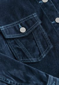 Donkerblauwe corduroy shirt met een borstzak met een zilveren knop, contrasterende lichte stiksels en een textuurafwerking.