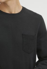 Black knit sweater featuring a round neckline, long sleeves, and a textured patch pocket on the left chest. Smooth fabric with a subtle sheen.