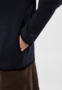 Námořnický pulovr s hladkou texturou a černými akcenty. Má boční kapsu a potištěné logo "BOSS" na rukávu.