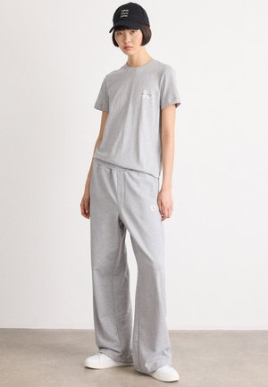 Grijze katoenen T-shirt met een logo op de borst, gecombineerd met losse grijze sweatpants met een elastische tailleband. Witte schoenen en een zwarte pet.