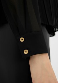 Blusa nera trasparente con maniche caratterizzate da una texture liscia, un design plissettato e due bottoni dorati sul polsino, con un tessuto nero sottostante.