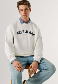 Witte katoenen trui met "PEPE JEANS" gedrukt in marineblauw. Gelaagd over een blauw geruit overhemd. Lichtblauwe spijkerbroek. Eenvoudig ontwerp.