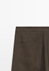 Donkerbruine stof met een achterzak en zichtbare bovenste stiksels, weergegeven tegen een witte achtergrond.
