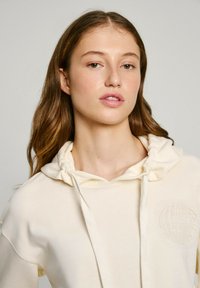 Sudadera color crema con una textura suave, cordones ajustables y un sutil logotipo en relieve en la parte frontal. Corte relajado y casual.