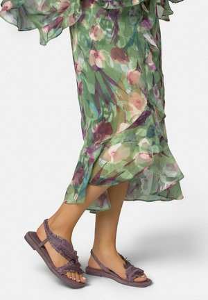 Pies con sandalias con cuentas color malva combinadas con un vestido verde floral transparente y fluido que termina a media pantorrilla.