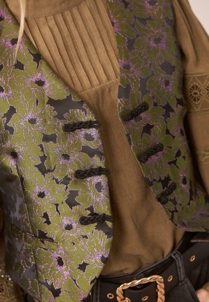 Chaleco floral verde con acentos morados, con un respaldo negro y cierres de toggle, superpuesto sobre una camiseta marrón de canalé. Detalles de tela con textura visibles.