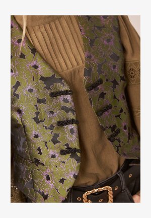 Chaleco floral verde con acentos morados, con un respaldo negro y cierres de toggle, superpuesto sobre una camiseta marrón de canalé. Detalles de tela con textura visibles.