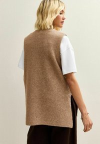 Bruine gebreide spencer met een geribbelde textuur, open zijkanten en een rechte snit, gedragen over een wit shirt met korte mouwen.