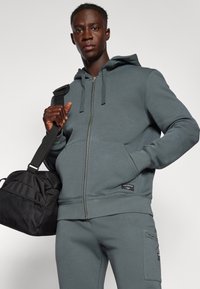 Sweatshirt cinza com fecho e material macio, com cordões, combinando com calças iguais. O modelo segura uma mala de desporto preta com detalhe em malha.
