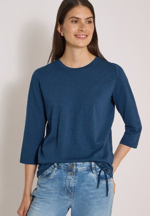 Femme vêtue d'un haut bleu à manches trois-quarts avec un détail noué sur le côté, associé à un jean en denim bleu clair, souriante et regardant légèrement vers la droite.