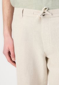 Světlé béžové lněné kalhoty s texturovaným povrchem, s páskem na šňůrku a bočními kapsami, kombinované s pomačkaným zeleným topem.