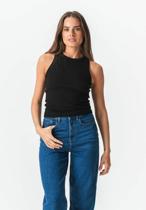 Fiatal nő hosszú barna hajjal, fekete ujjatlan felsőt és magas derekú kék farmert visel, egy kezét a zsebében tartva áll.