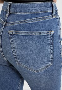 Modré džíny z denimu s vysokým pasem, vybavené zadní kapsou a texturovaným povrchem. Švy jsou viditelné podél švů.