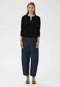 Svart ribbad cardigan med rund halsringning och fem knappar, i kombination med löst sittande mörkblå jeansbyxor och svarta flip-flops.