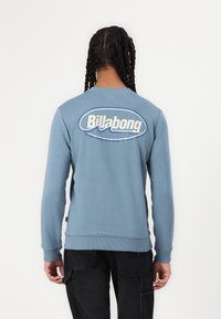 Blå crewneck sweatshirt med lange ermer, laget av myk stoff. Har en stor hvit Billabong-logo på baksiden. Ribbestrikkede mansjetter og nederkant.