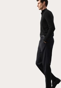Černý svetr se zipem u krku spárovaný s tmavými kalhotami. Tkanina vypadá hladce, má přilnavý design a minimalistické detaily.